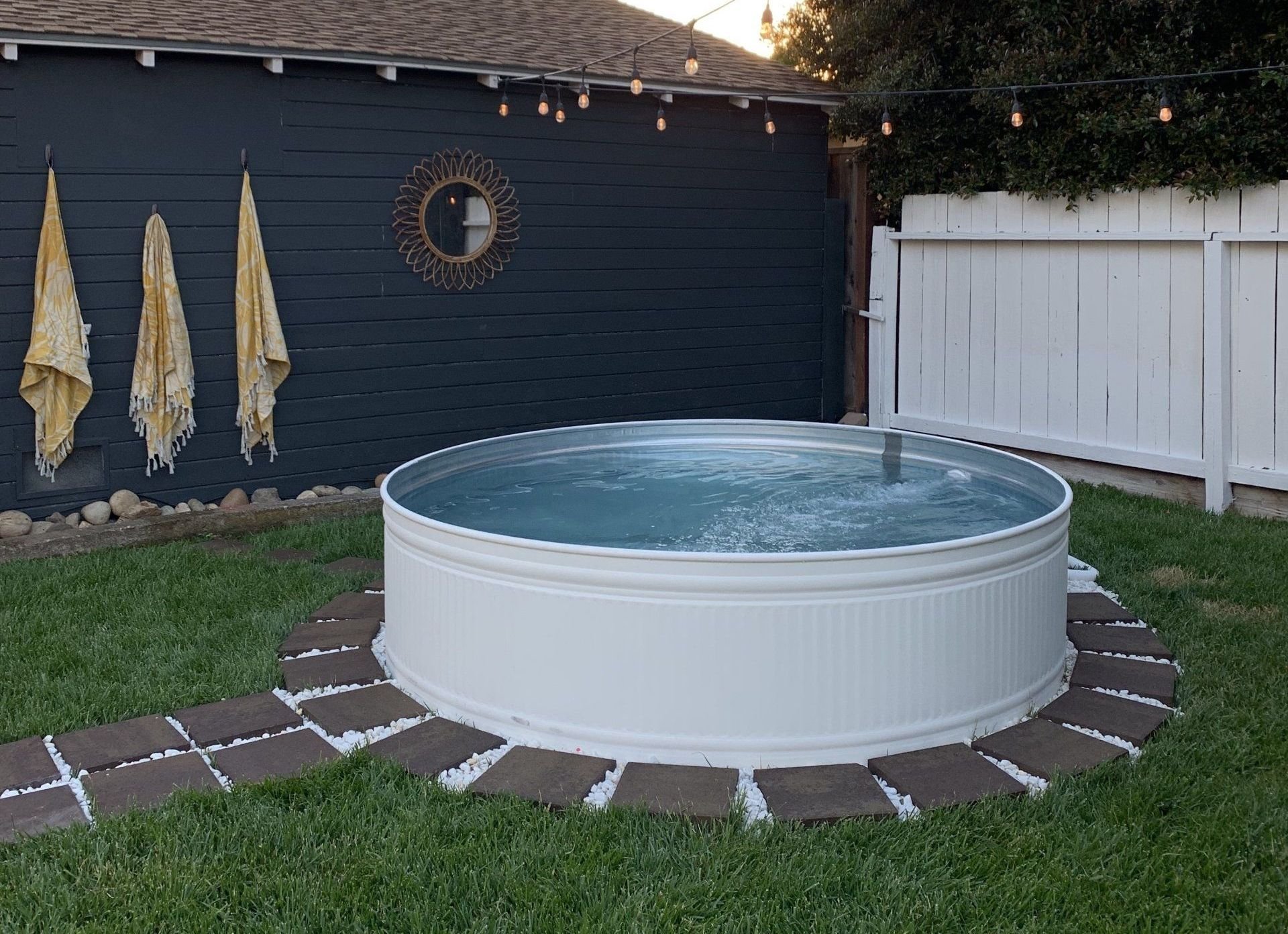 A traditional style backyard with a stock tank pool painted white surrounded by green lawn and overhead string lights
