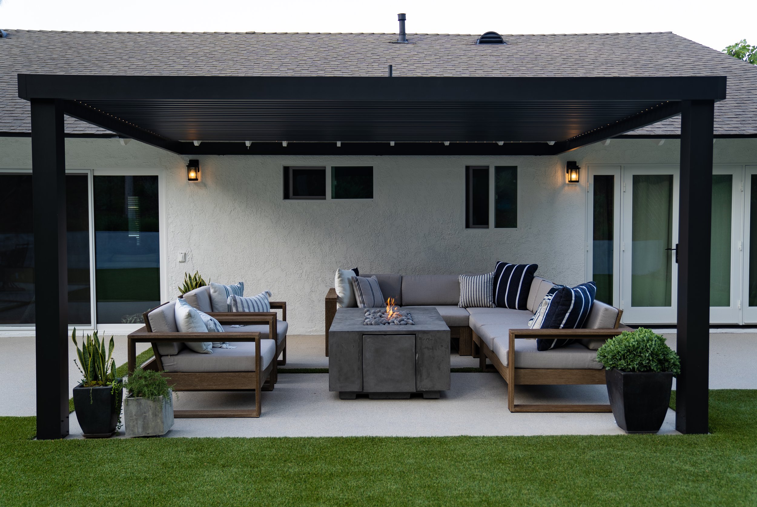 black pergola attached to back of home over a fire pit seating area with sectional and two lounge chairs